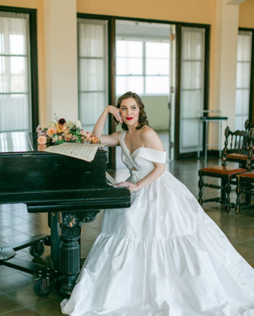 The Bride in ballgown from plain white fabric, classic Hollywood waves and red lipstick, sitting by the piano