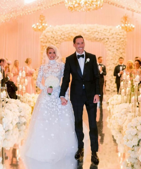 Paris Hilton wedding, white rose flower arrangements, shiny floor, white venue, black tuxedo
