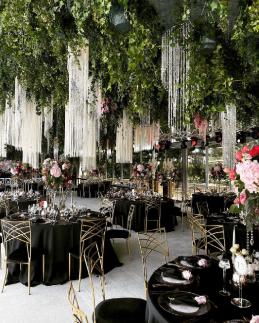 Wedding venue with round tables covered with black table cloths, black plates, gold metal chairs, roof with leaves and string crystal chandeliers, pink flower bouquets in high vases