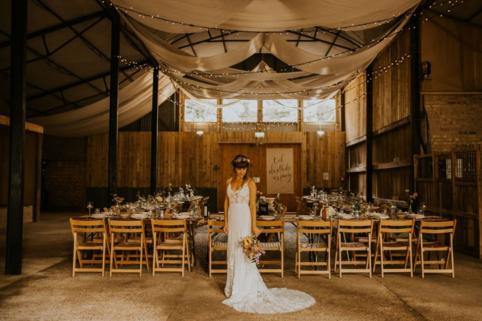 Small barn wedding reception tables, the Bride in rustic airy gown with woven on her head and meadow flowers bouquets, high ceiling with string lights and white fabric
