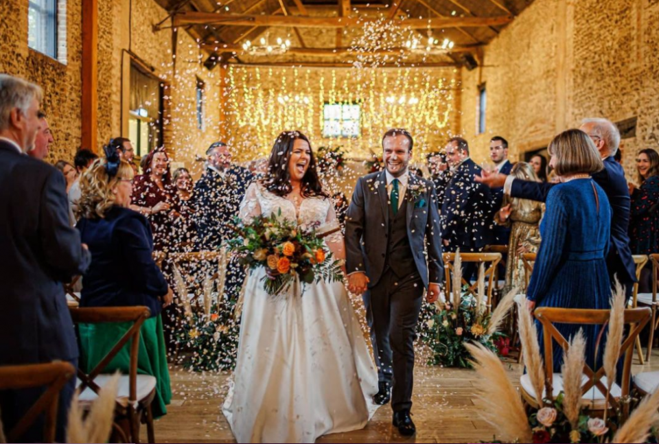 Stone barn wedding, newly-weds after the ceremony, guests throwing rice at them, high ceiling and chandeliers, visible wooden construction of the roof 