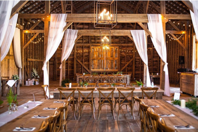 Wooden barn decorated with string lights, white fabric and subtle greenery. Geometrical chandeliers with candle-looking bulbs. Wooden reception tables and chairs