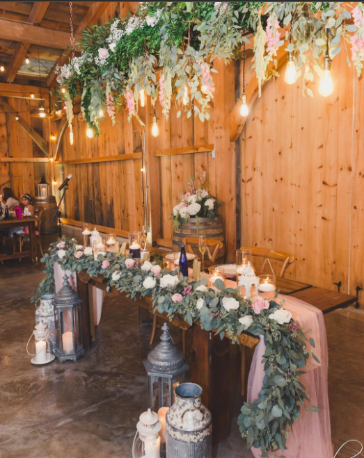 Sweetheart table decorated with pink chiffon and pastel coloured flower arrangements on the table and hanging above from the ceiling. Old lanterns and milk cans on the floor