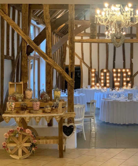White walls barn with visible wooden construction elements, tiles on the floor, sweets cart, wedding round reception tables and LED light LOVE sign