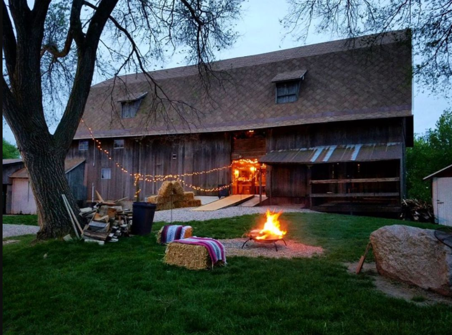Classic barn with bonfire in the front and pillow and hay bales to chill out, string lights in front of the building hanging from the tree