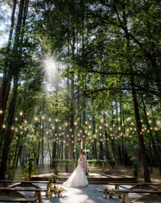 Forest wedding venue with string lights hang between trees and wedding couple standing in the daylight