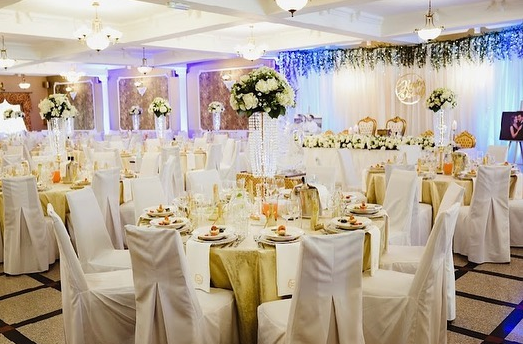 White and golden colored venue, chairs covered with white cloth, white bouquets in high vases, sweetheart table with white roses