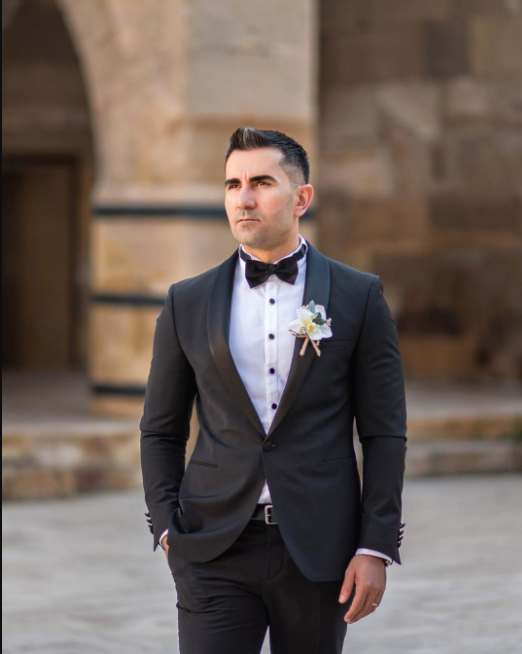 The Groom in classic black suit with bow tie and small bouquet pinned to the suit, black buttons, black hair in fashionable hairstyle