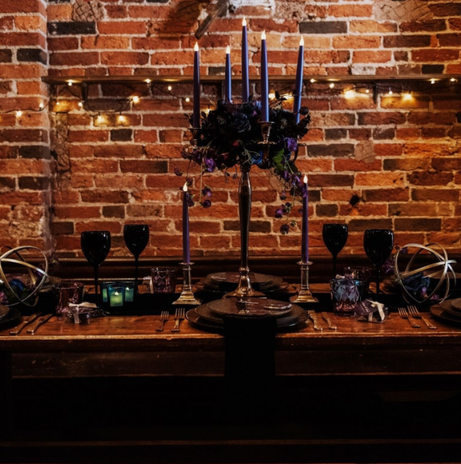 Candelabra with long dark blue candles, high candleholders with purple candles, black plates and wine glasses, blue juice glasses, brick wall on the background