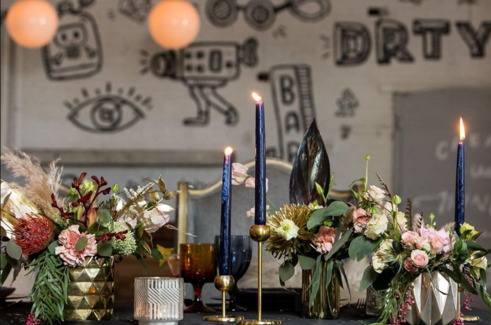 Golden and pastel bouquets, long holographic blue candles in golden candleholders, wall covered in simple graffiti