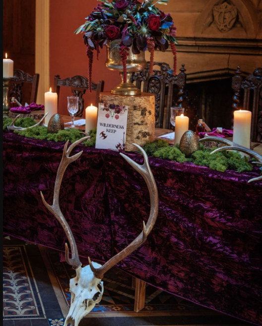 Purple velvet tablecloth, deer skull on the floor, big white candles, tree stump under the golden vase with dark-colored bouquet, vintage chairs with ornamented wood
