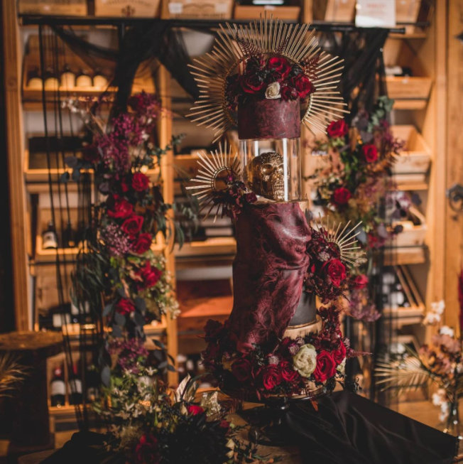 Raspberry and black colored 3 levels wedding cake with golden crown on top and golden skull, decorated with ecru and red roses