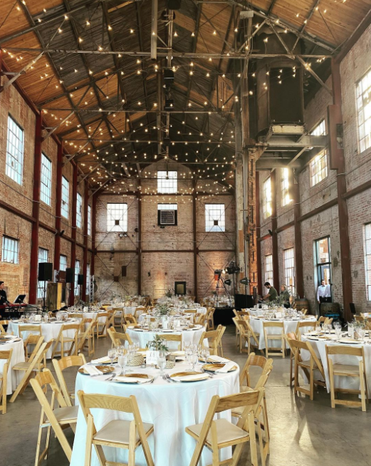 High ceiling, red brick venue with visible wood construction, round tables with light wooden chairs