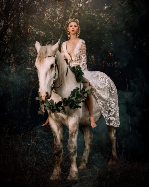 Blond bride in laced wedding dress on the white horse decorated with leaves, deep dark forest in the back