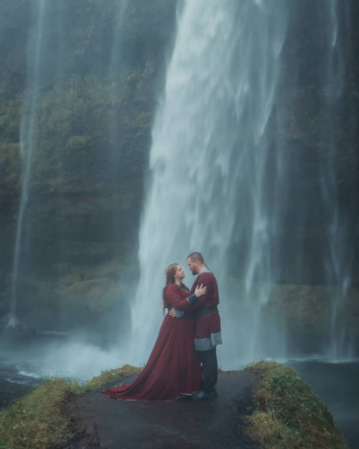 Couple in deep, dark red costumes under the misty waterfall standing on the rock