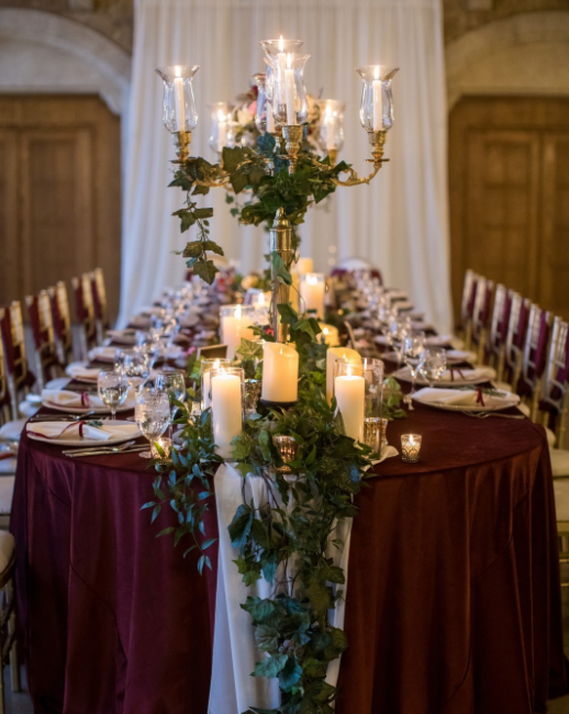 Golden candelabra decorated with leaves, white thick candles, long table covered in dark red tablecloth