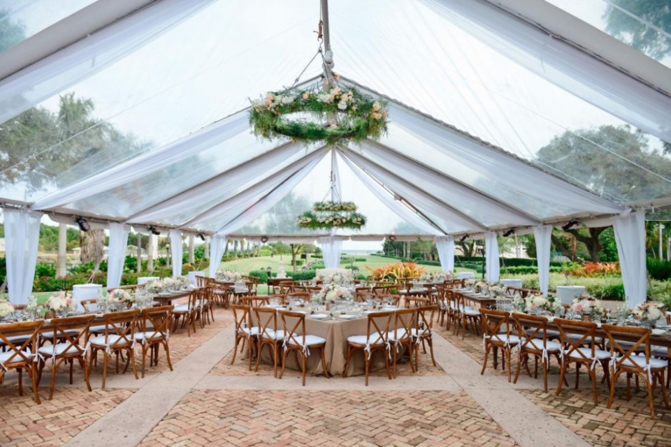 Wedding marque with chandeliers with greenery, beige table cloths and white chair pillows, huge garden
