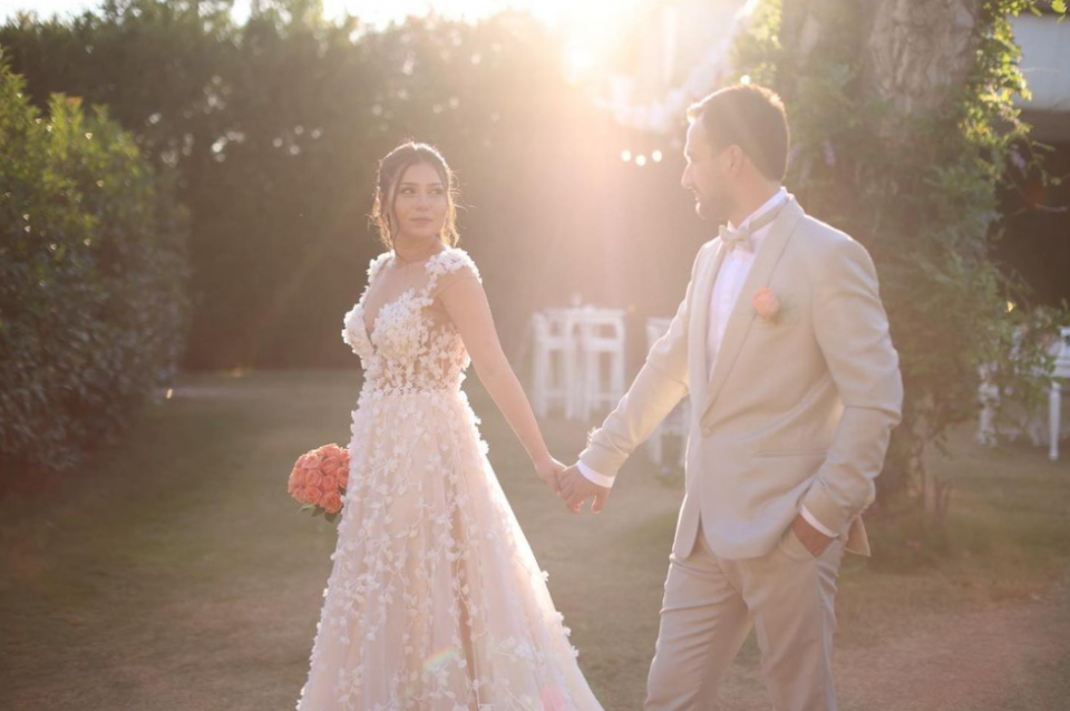 Wedding couple, groom in light suit with salmon colored rose, bride in dress with 3d flower applications and salmon colored rose wedding bouquet. Standing in the sunshine in the garden