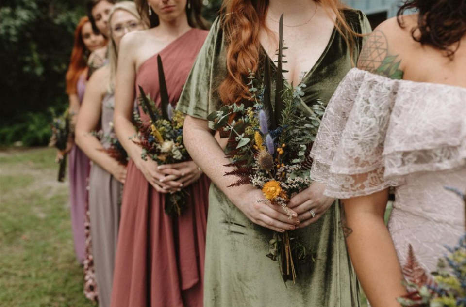 Bride in laced wedding gown and bridesmaids standing behind her in linen gown, each in different color holding bouquets made out of dried greenery